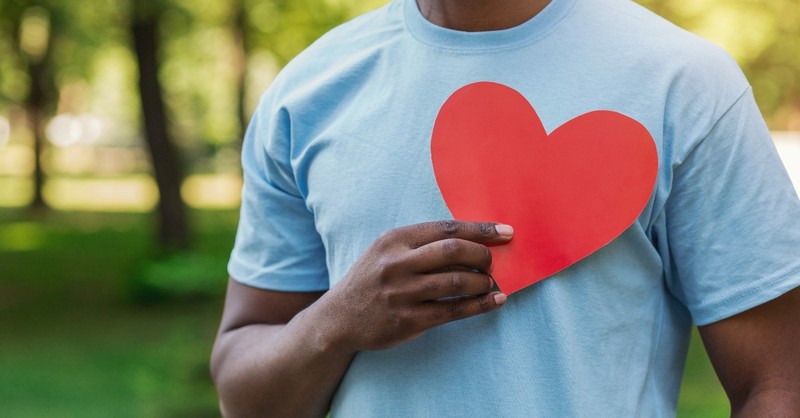 man in blue t shirt holding up cut out red heart in front of his chest, prayers to check heart