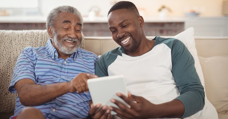 grown son with senior dad laughing on couch with ipad Fathers Day