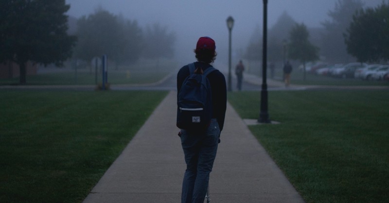 young man with backpack standing in dark fog on college campus