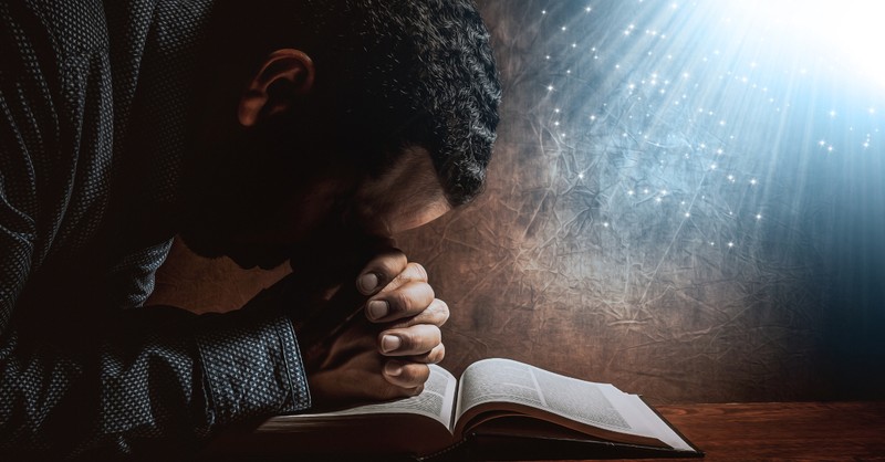 man resting head on praying hands on open bible light streaming above holy spirit, prayers for hidden strength