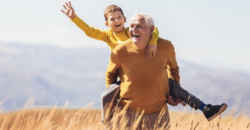 senior grandparent giving grandson a piggyback outdoors in golden field