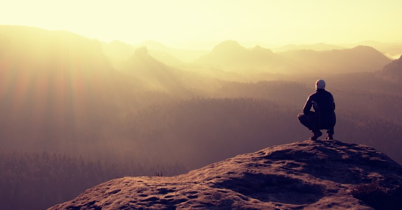 man crouching on mountain top sun setting