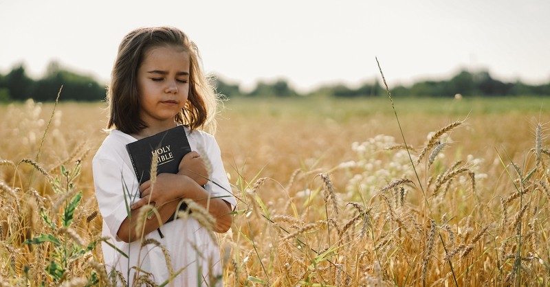 little girl holding Bible outside in a field with her eyes closed, Bible verses for kids