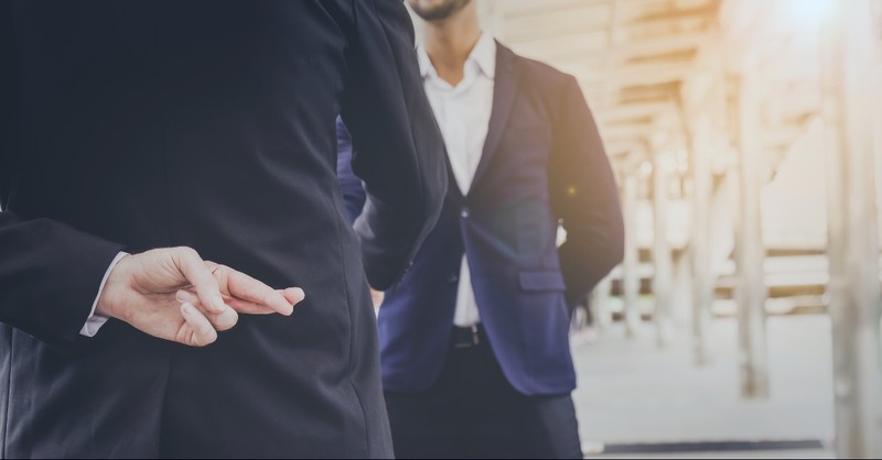 Man shaking another man's hand with crossed fingers behind his back