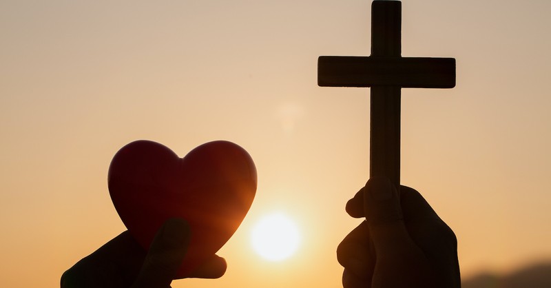silhouette of hands holding up a heart and a cross outside at sunset, love your enemies