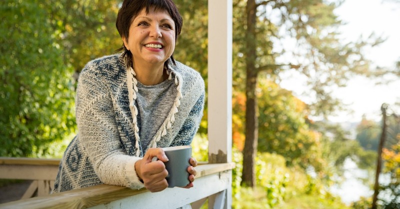 mature woman standing with mug on front porch in autumn looking relaxed and happy, tips for simple stress-free fall