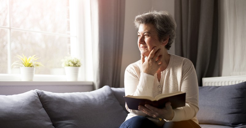 older woman sitting in living room with Bible thinking toward window