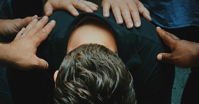 Man sitting with head bent, others laying hands on him in prayer