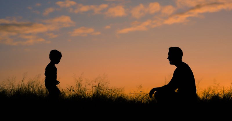 silhouette of dad sitting in grass waiting for toddler son to walk to him