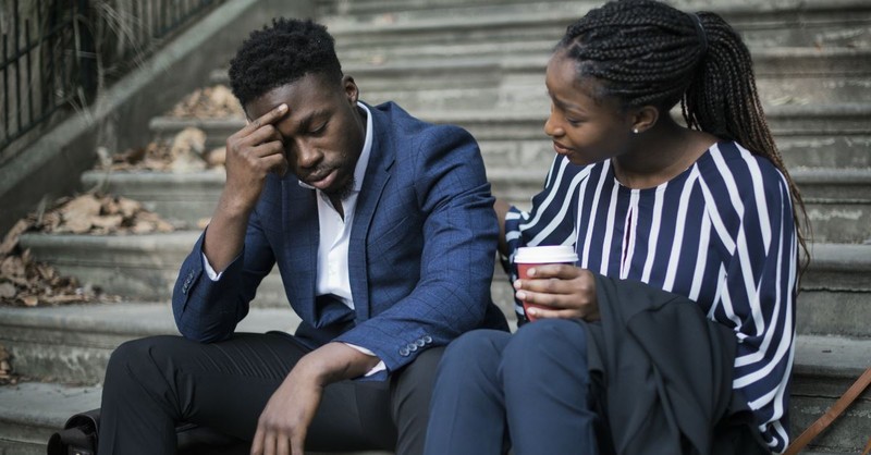 woman comforting man who looks distressed, both in business clothes sitting on steps