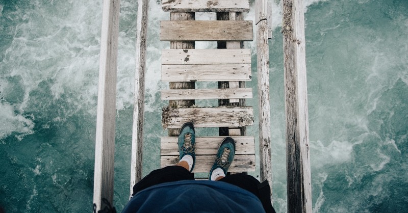 down view of feet about to cross weathered open-slat footbridge over water