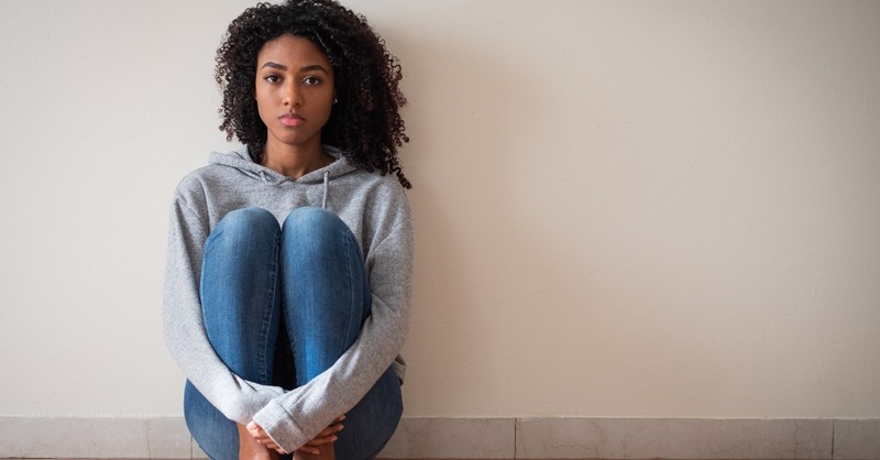 worried woman sits against a wall, her arms wrapped around her legs