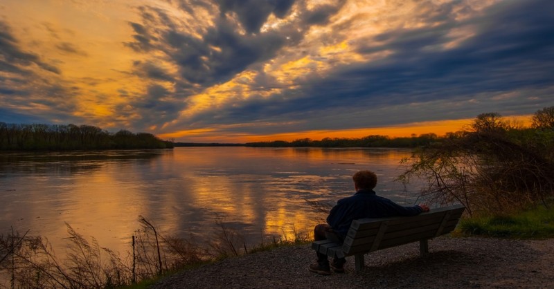 Older man sitting on wooden bench by Missouri River looking at dramatic sunset