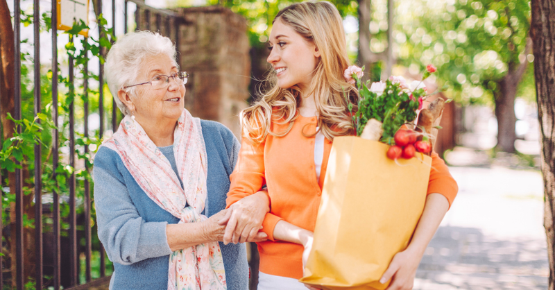 Younger woman humbly helping an elderly woman carry her groceries home.