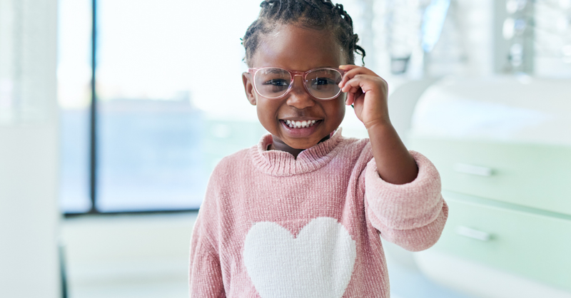 Happy little girl with glasses and a pink sweater with a white heart on it; a prayer for our children to know God's love.