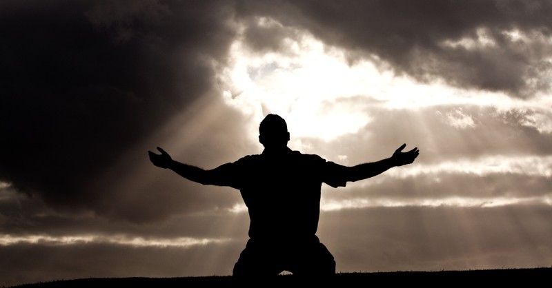 Silhouette of a man kneeling with arms raised towards the light in praise