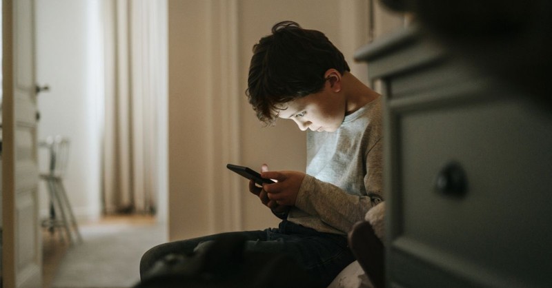 Young boy on a smartphone in a dark room; the solution to minors' exposure to porn, 'Keeping unsupervised tech out of unsupervised hands.'
