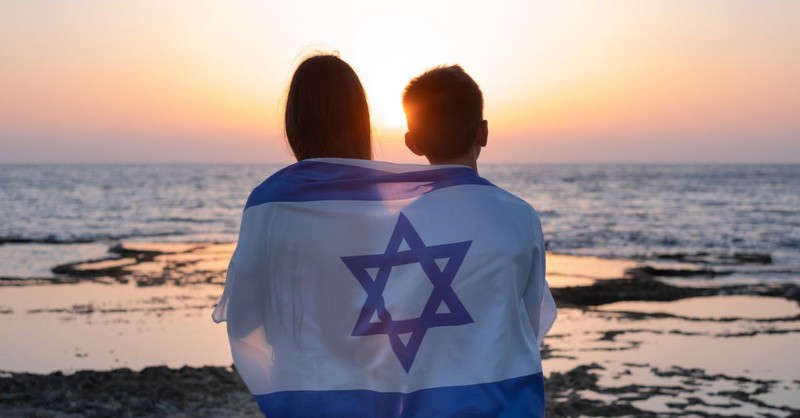 Teens sitting on a beach in Israel with the Israeli flag wrapped around them.