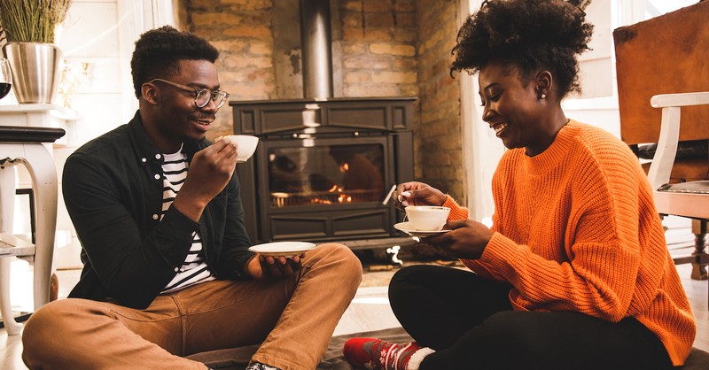 Cute couple laughing drinking tea cozy by fireplace in winter