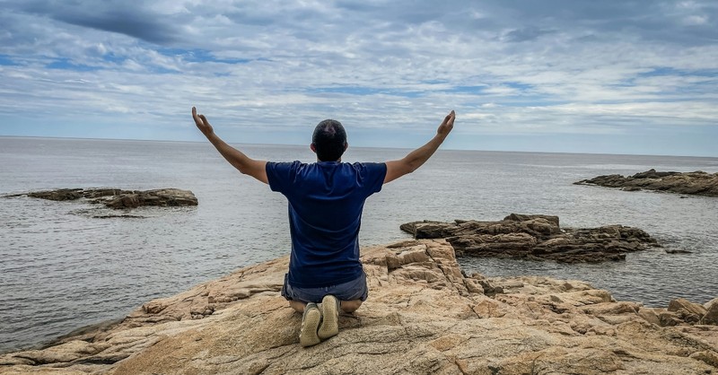 A man kneeling by the ocean with arms raised in worship