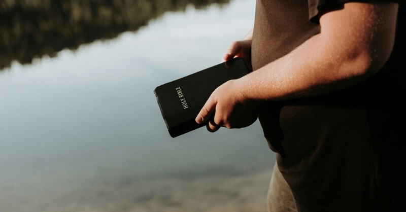 **Alt Text:** A person stands beside a calm body of water holding a Bible, reflecting on faith, Scripture, and personal devotion in a peaceful outdoor setting.