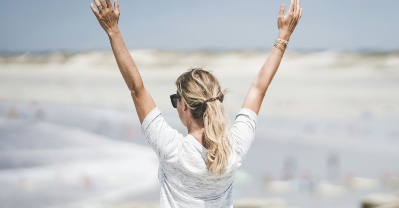 A woman stands outdoors with her arms raised toward the sky, expressing gratitude, praise, and joyful worship in an open natural setting.