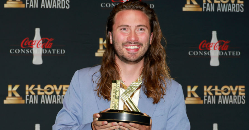 Christian music artist Brandon Lake smiles while holding a K-LOVE Fan Award trophy at the K-LOVE Fan Awards, standing in front of the event backdrop.