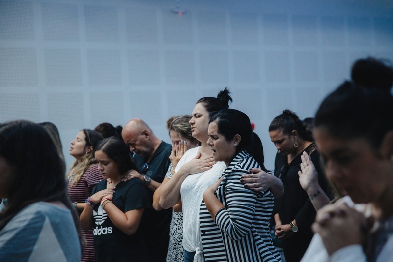 At church meetings, different groups of Christians stand together and pray, many with their eyes closed and hands on their chests or leaning on each other's shoulders, expressing worship, unity, and intercession.