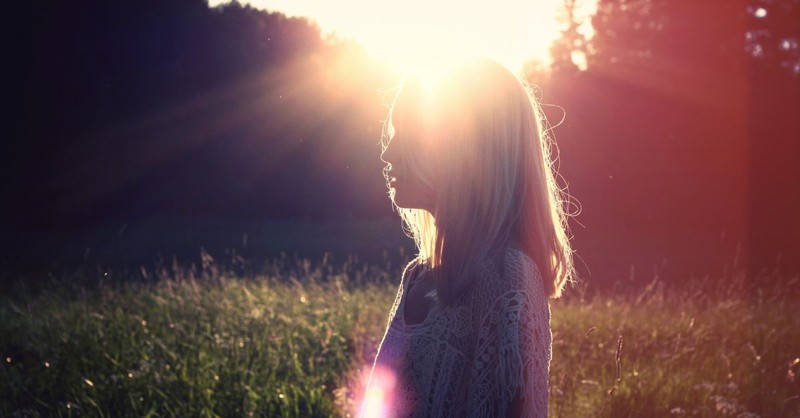 A peaceful photograph of a woman standing in a sunlit field at golden hour, her face turned toward the warm light as the sun shines behind her, symbolizing hope, renewal, God's presence, and the light of Christ breaking through the darkness.