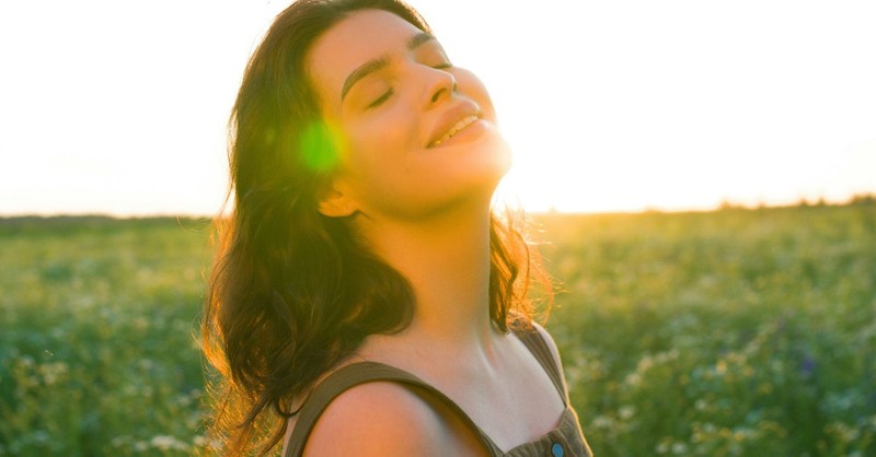 A joyful young woman stands in a sunlit field with her eyes closed and face lifted toward the warm evening light, expressing peace, gratitude, freedom, and the joy of resting in God's presence.