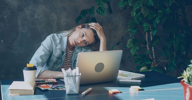 A woman sits at her desk with her head resting in her hand as she looks at her laptop, surrounded by notebooks and work supplies, conveying stress, mental fatigue, or burnout while working from home.