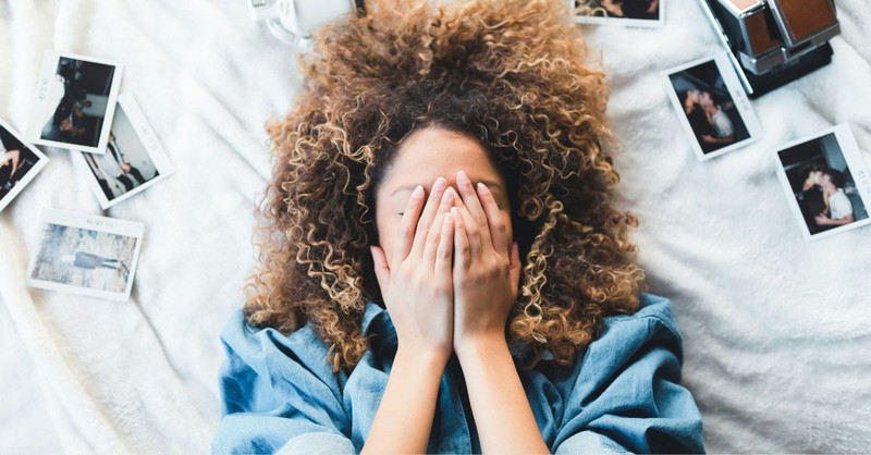 A woman lies on a bed with her face covered by her hands, surrounded by instant photos and a camera, expressing grief, regret, heartbreak, or emotional pain while reflecting on difficult memories.