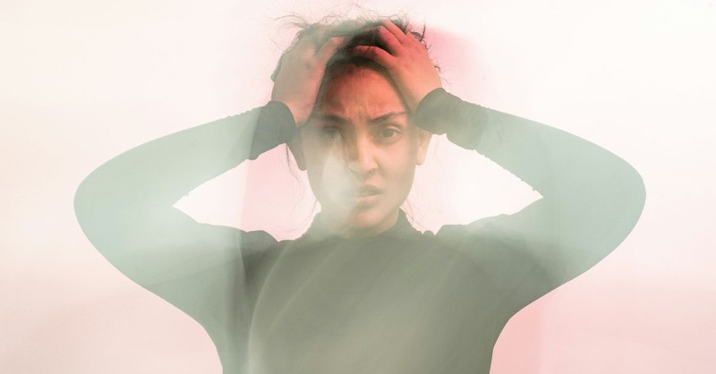 A woman grips her head with both hands and looks forward with a distressed expression as soft light and haze surround her, illustrating anxiety, mental overwhelm, panic, or emotional turmoil.