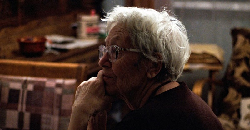 An elderly woman with white hair and glasses rests her chin on her hand while sitting in a warmly lit room, appearing thoughtful and reflective as she looks toward someone out of frame.