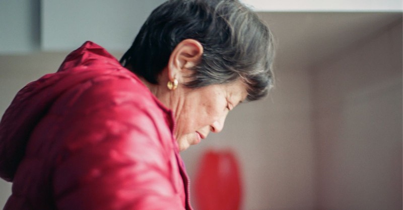 An older woman with short gray hair bows her head with her eyes closed while wearing a red jacket, conveying a quiet moment of prayer, grief, reflection, or trust in God during a difficult season.
