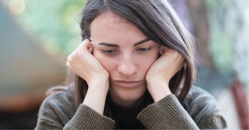A young woman rests her face in her hands and looks down with a discouraged, weary expression, conveying sadness, emotional exhaustion, anxiety, or discouragement as she quietly reflects.
