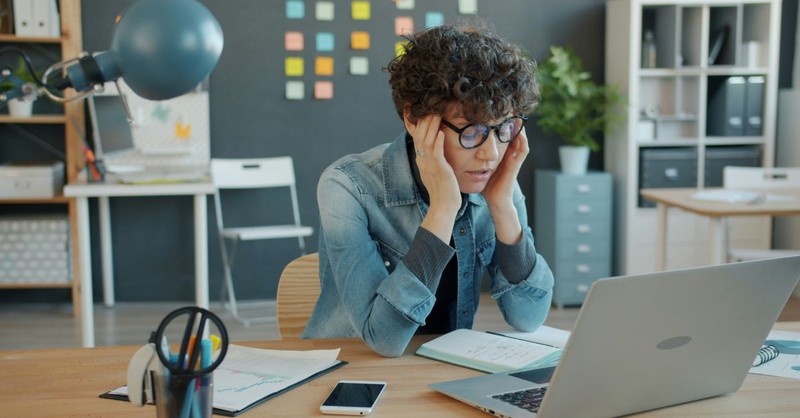 A woman sits at a desk holding her temples while staring at a laptop in a modern office, surrounded by notebooks and work materials, conveying stress, mental overload, decision fatigue, or workplace anxiety.