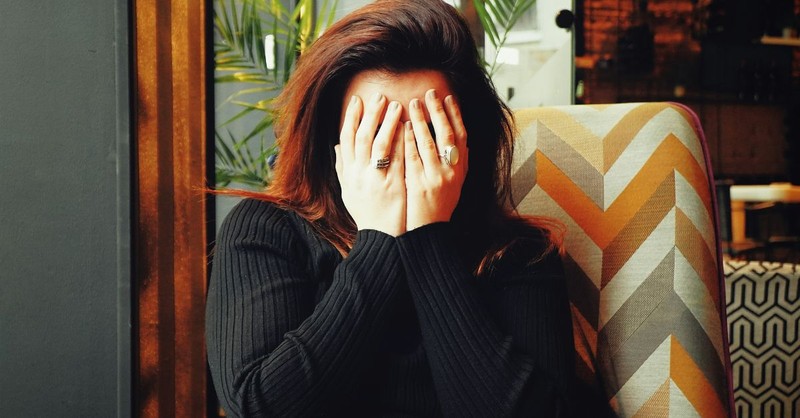 A woman sits indoors with her face buried in her hands, conveying deep emotional distress, grief, anxiety, or overwhelm as she seeks a moment of privacy amid a difficult situation.