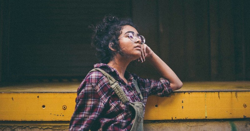 A young woman wearing glasses and casual clothing leans on a railing while gazing thoughtfully into the distance, conveying contemplation, waiting, discernment, or seeking God's direction for the future.