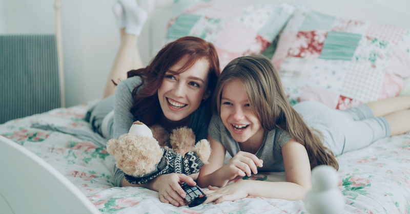 A mother and daughter laugh together while lying on a bed at home, symbolizing the joy of a close parent-child relationship, intentional connection, and nurturing faith within the family.