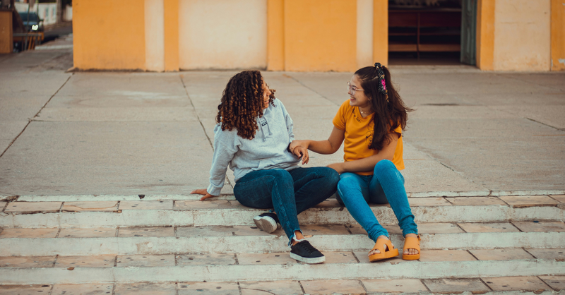 Two girls sit together on outdoor steps, smiling and talking, symbolizing friendship, encouragement, and the importance of building confidence, kindness, and faith in the next generation.