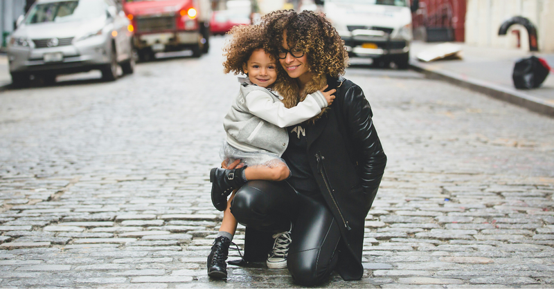 Mother holding her young daughter as they smile and embrace together on a city street.