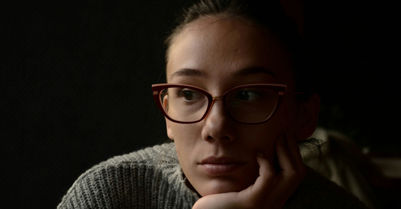 Thoughtful woman wearing glasses rests her face on her hand while looking to the side in a quiet, contemplative moment.
