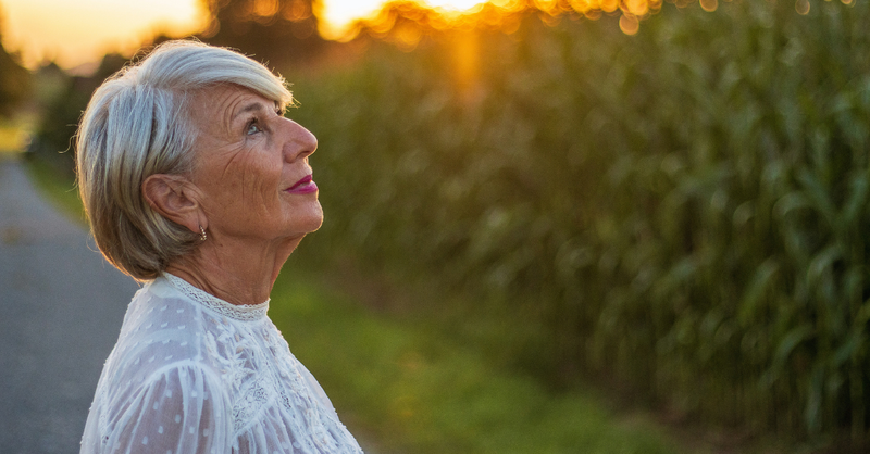 Older woman with gray hair looks upward in quiet reflection outdoors as warm sunlight glows behind her.
