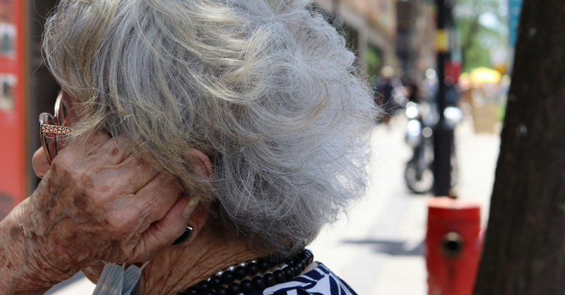 Older woman with gray hair and glasses holds her hand near her ear while standing on a busy city sidewalk.