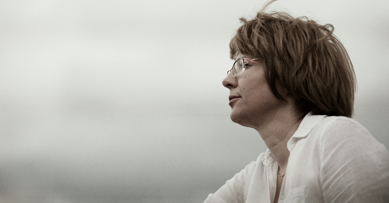 Thoughtful woman wearing glasses looks into the distance against a muted gray sky.