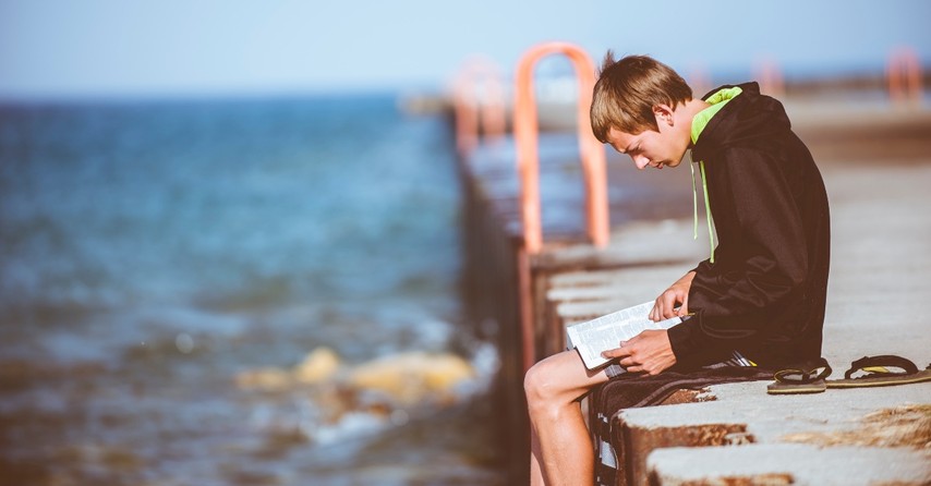 Teenage boy reading the Bible while sitting on a dock