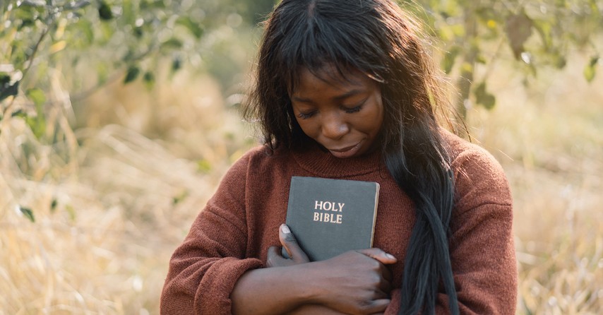 woman holding Bible outside in nature with head bowed in prayer, what is faith in Hebrews 11