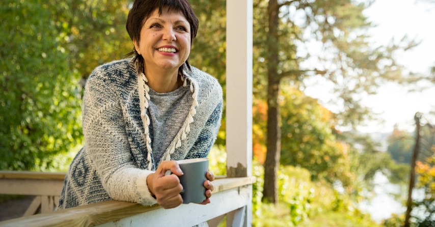 mature woman standing with mug on front porch in autumn looking relaxed and happy