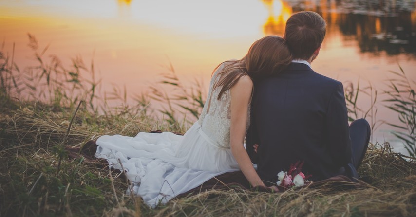 bride and groom sitting by lake at sunset